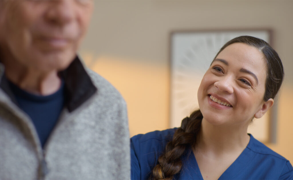 A healthcare professional in blue scrubs smiles warmly at an elderly person wearing a gray sweater, embodying the principles of how to be a caregiver.