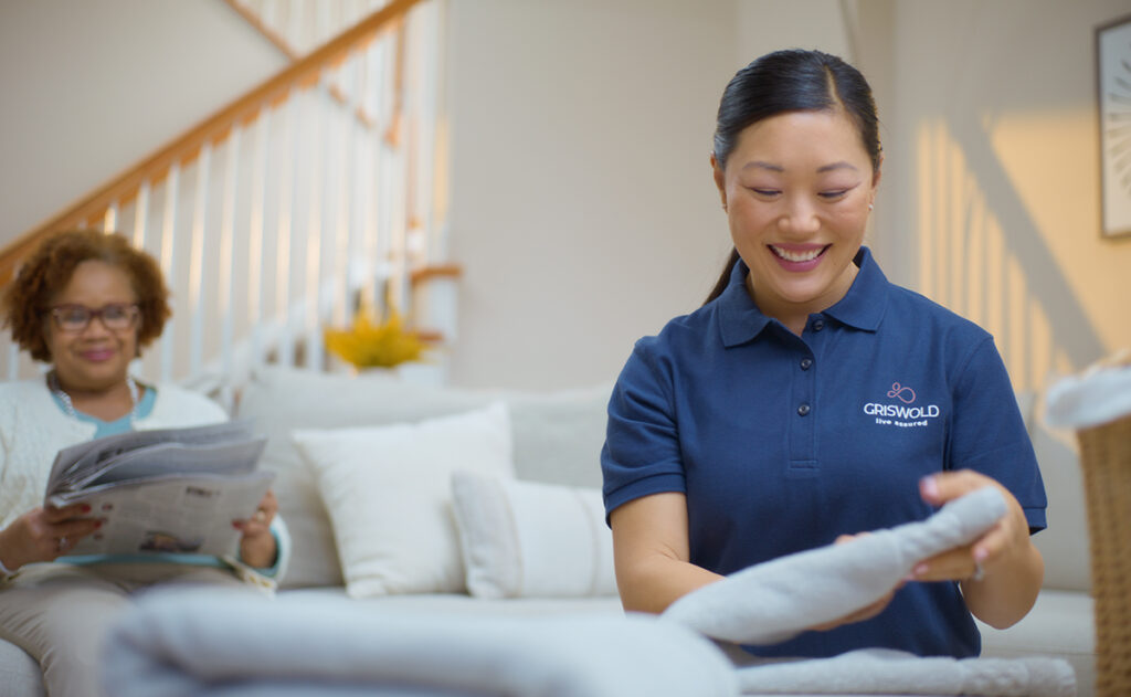 A woman in a blue uniform labeled "Griswold Home Care" folds laundry, showcasing homemaking services, while another woman reads a newspaper on the couch in the background.