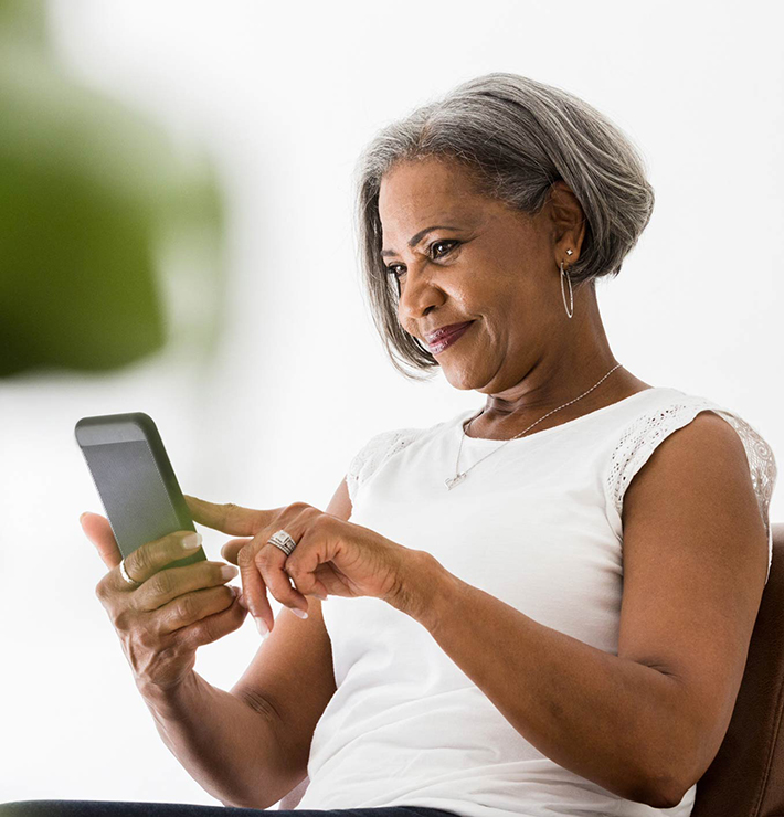 An older woman with short gray hair, wearing a white shirt, sits and uses a smartphone, smiling slightly and tapping the screen with her index finger.