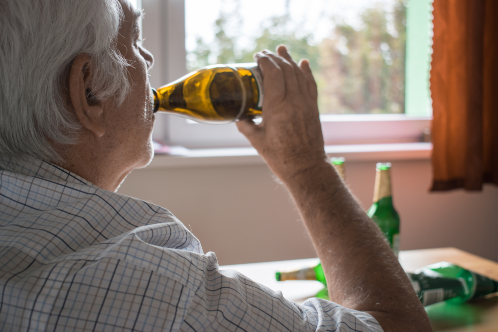 An older man drinks from a beer bottle while sitting indoors