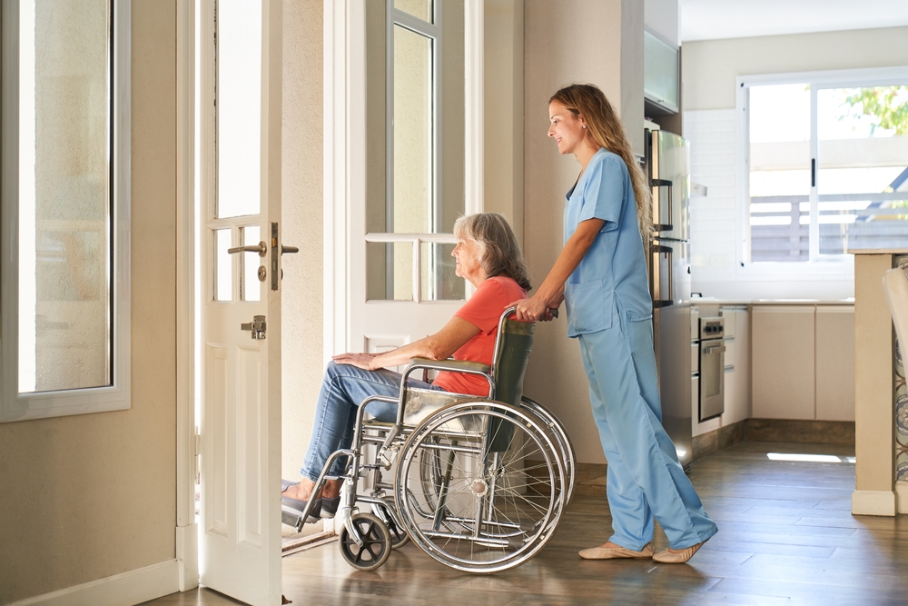 A healthcare worker pushes an elderly woman in a wheelchair towards a sunlit door in a home setting.