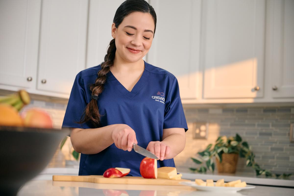 A person in a blue uniform is slicing an apple on a wooden cutting board in a bright kitchen.