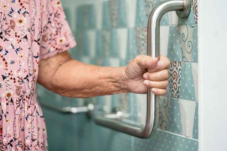 An elderly person wearing a floral dress holds onto a metal grab bar installed on a bathroom wall with patterned tiles.