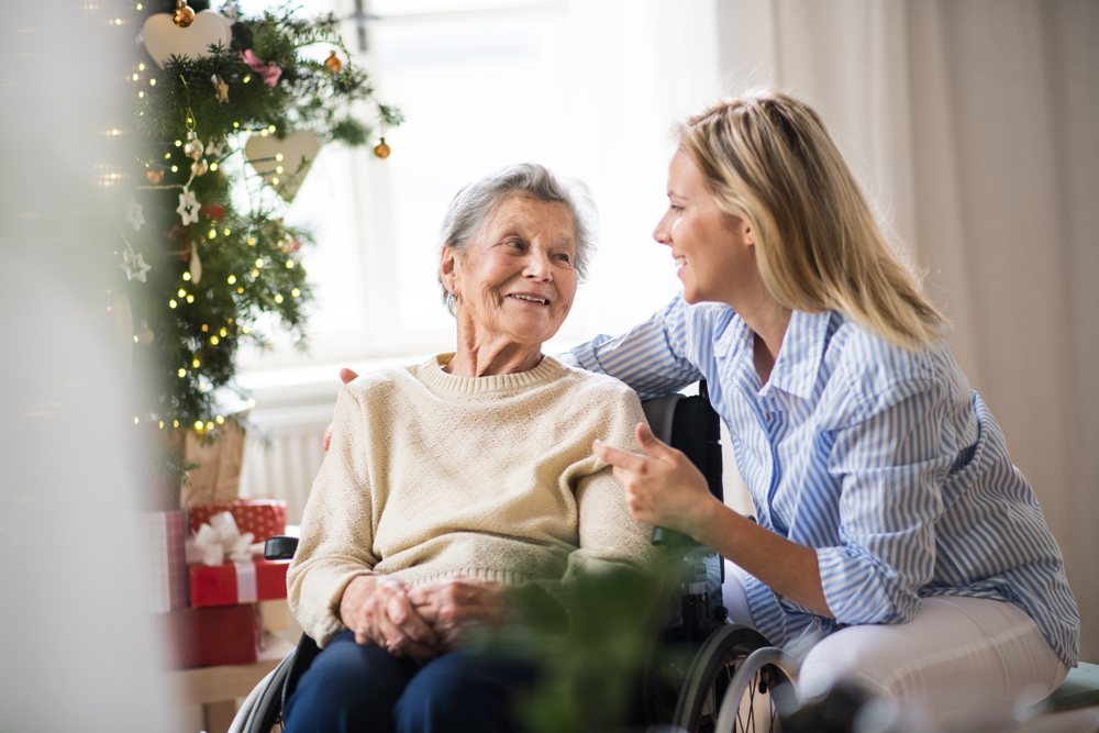 An elderly woman sitting in a wheelchair smiles at a younger woman in a cozy room with a decorated Christmas tree in the background.