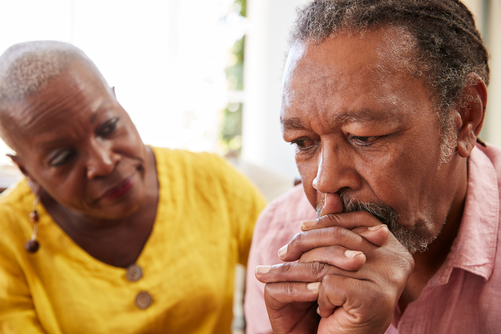 An elderly man appears deep in thought while a woman gently places her hand on his shoulder, offering comfort.