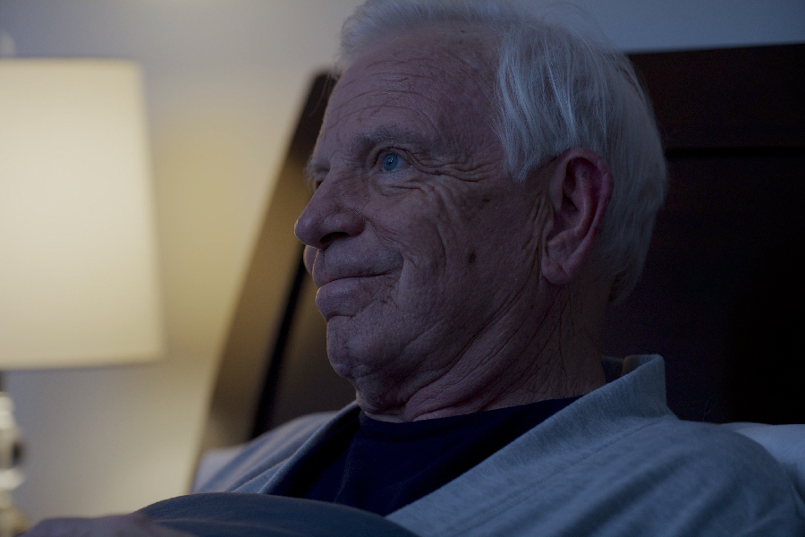 Elderly man with white hair and a neutral expression sitting in bed, illuminated by a bedside lamp.