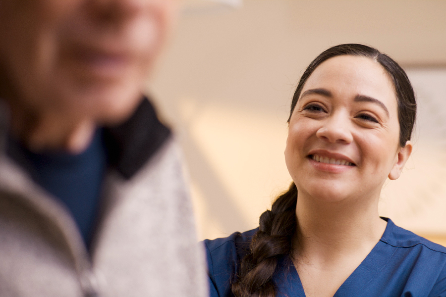 A smiling woman in a blue uniform looks at an older man in the foreground, who is facing away.