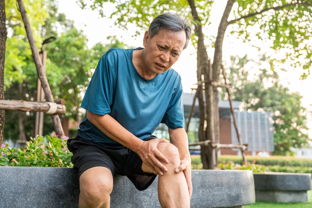 An older man sitting on a bench in a park holds his knee with a pained expression, suggesting discomfort or injury.