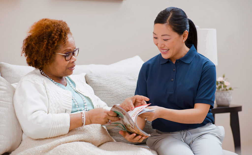 A woman sits on a couch with a caregiver. The caregiver smiles and points to a page in a magazine while the woman looks at it.