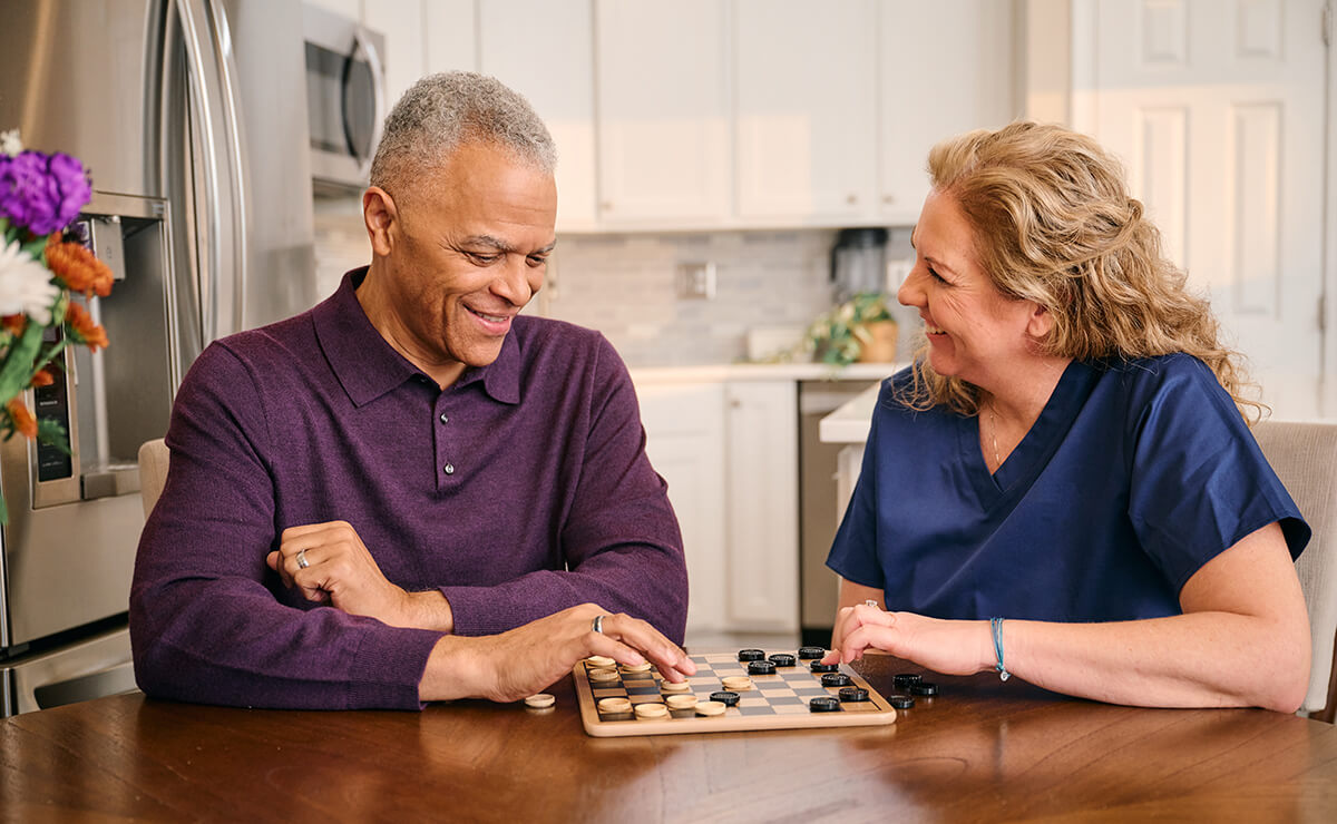 A man and a woman play checkers at a kitchen table. The man is wearing a purple sweater, and the woman is in blue scrubs. They are smiling and engaged in the game.