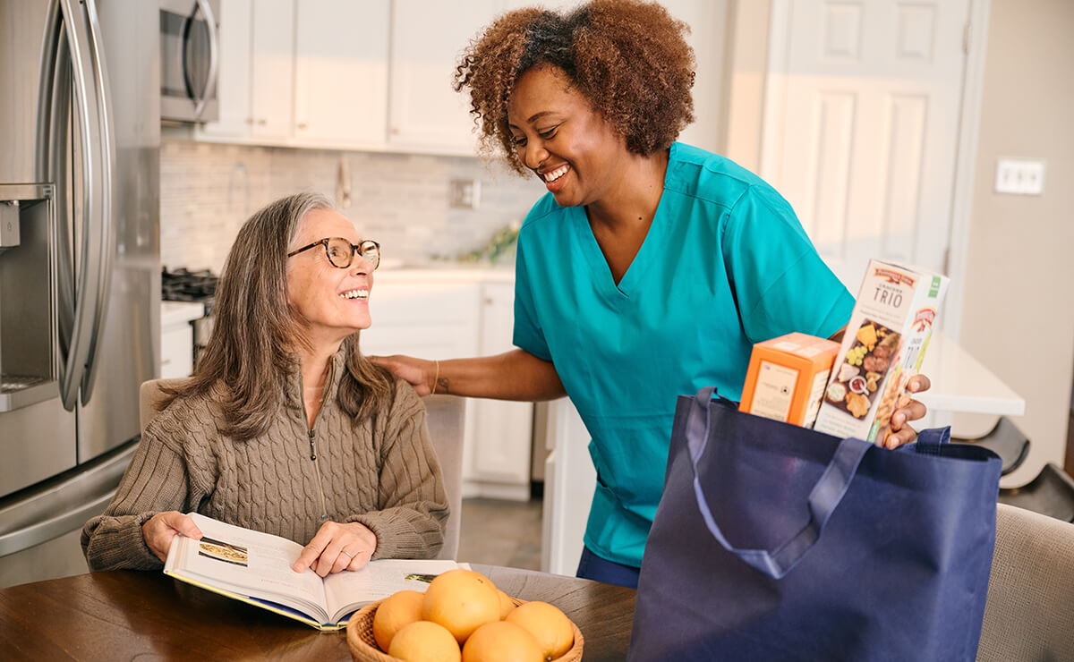 A caregiver in blue scrubs smiles at an elderly woman with glasses who is sitting at a table with a book and bowl of oranges. A bag of groceries sits on the table.