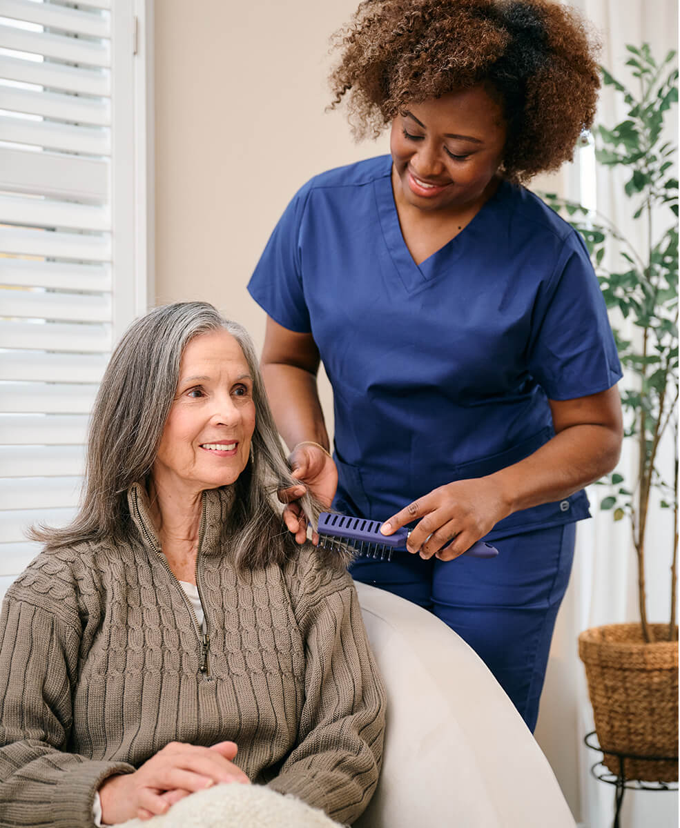 A caregiver in blue scrubs assists an elderly woman by combing her hair. The elderly woman is sitting and smiling. A plant and window blinds are in the background.