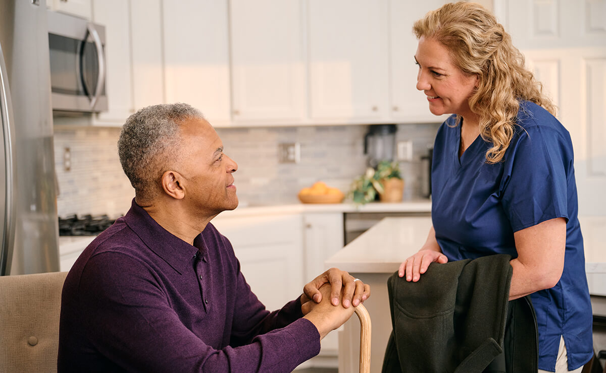 A man sits at a table, smiling and holding hands with a woman in blue scrubs, who is standing and looking at him. They are in a kitchen setting.